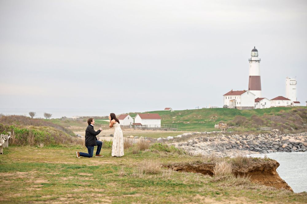 Romantic marriage proposal at Montauk Point Lighthouse, man kneeling before woman on grassy coastal cliff, New York.