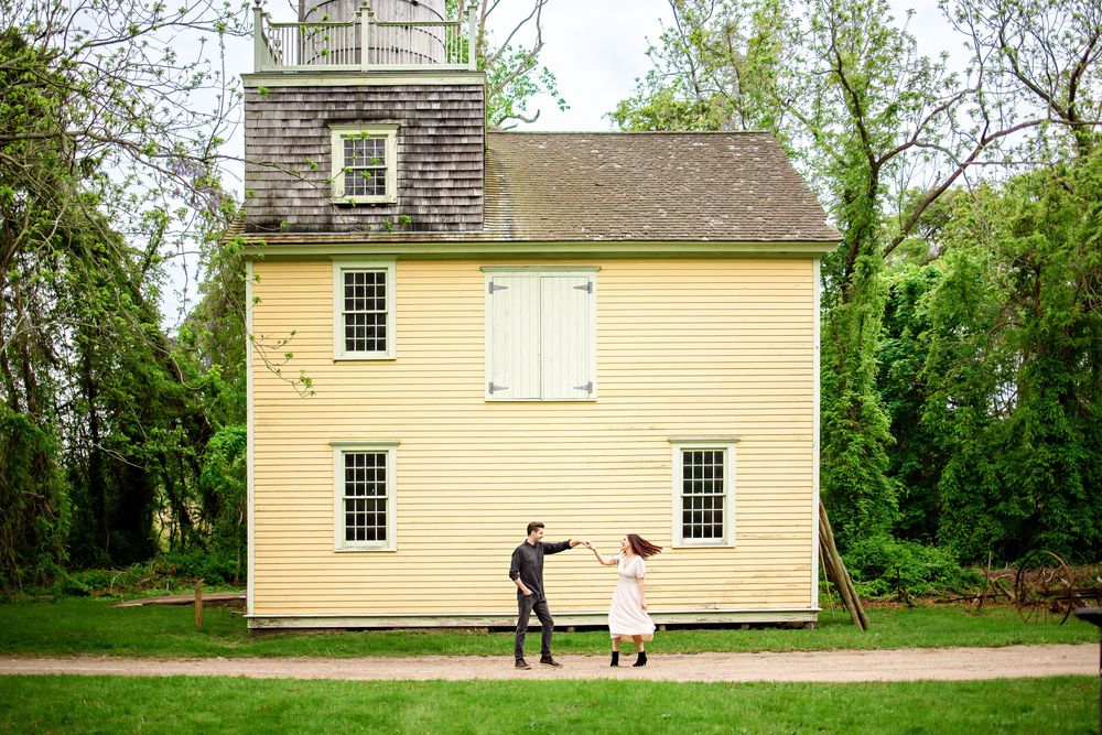 Two figures in formal attire dance playfully in front of a historic yellow colonial house with a square tower.