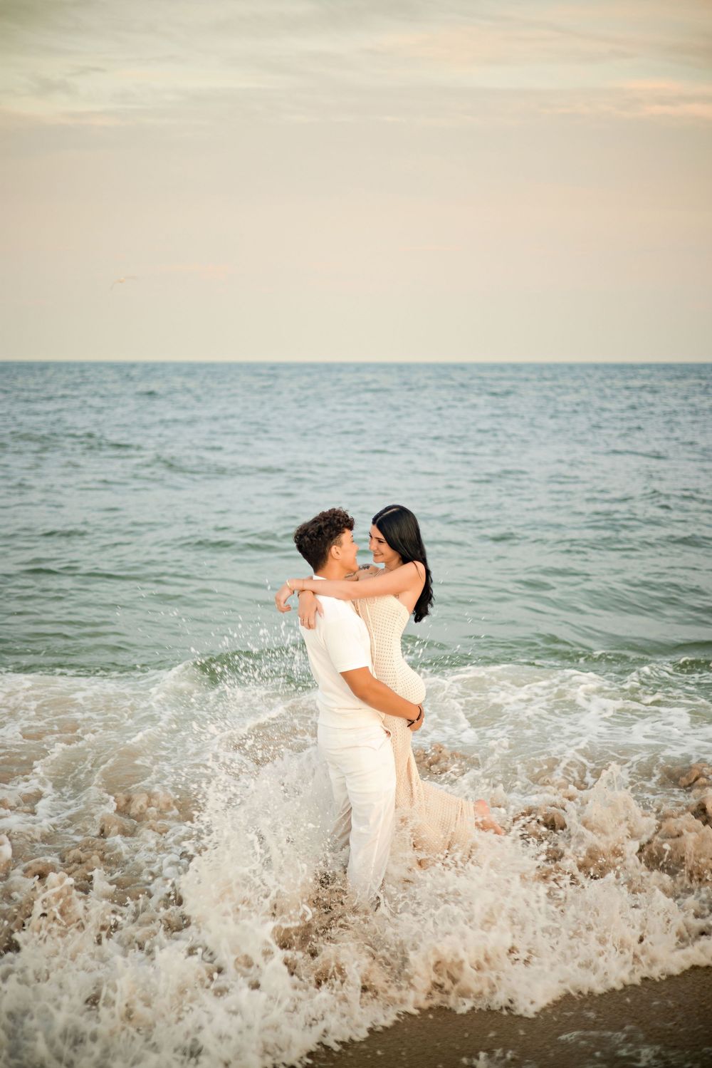 Romantic couple embracing in ocean waves on a beach, both dressed in white, sharing a tender moment at sunset.