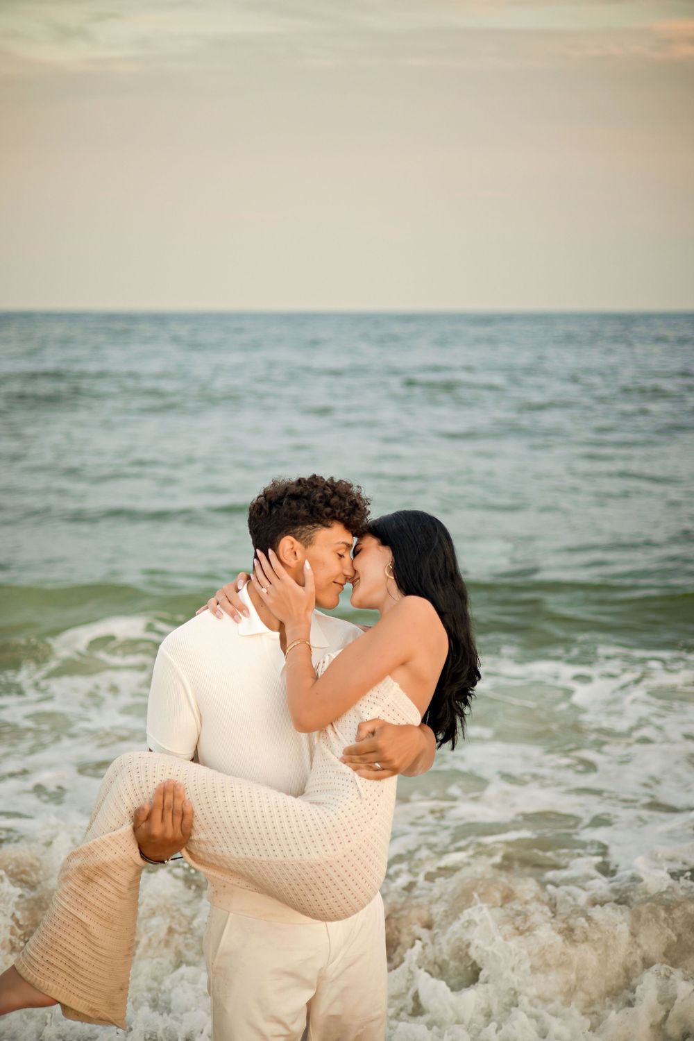 Romantic couple kissing on a beach at sunset, man carrying woman in white outfits with ocean waves behind them.