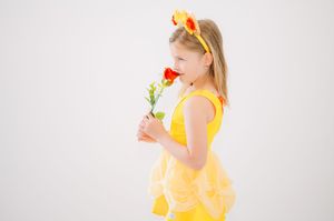 Young girl in yellow dress holding single red rose flower stands in profile against white background.