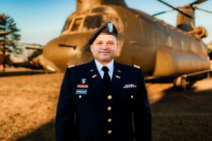 Military service member in formal uniform standing proudly in front of a CH-47 Chinook helicopter at sunset.