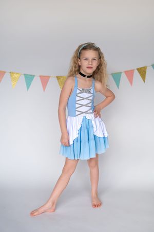 A child poses in a light blue and white dress with laced bodice against a gray background with colorful bunting.