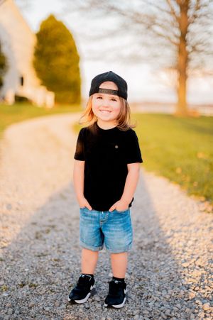 Young child wearing baseball cap and casual outfit standing on gravel path.