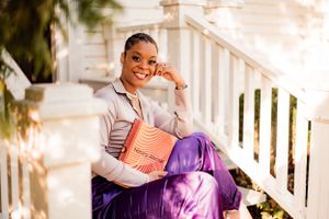 Person in casual attire sitting on white porch steps holding an orange book.