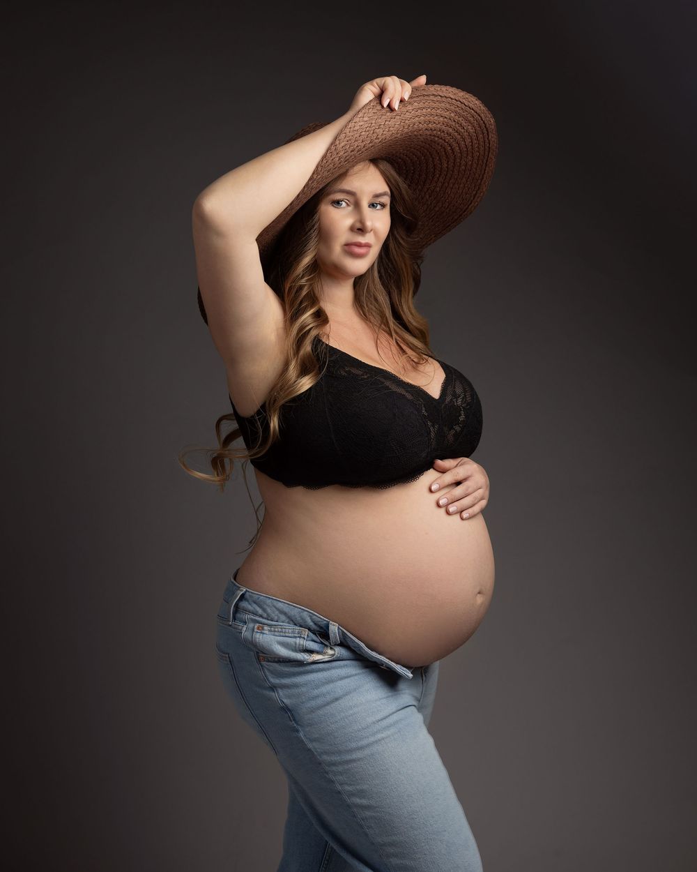 Pregnant woman in black lace bra and jeans holding a wide-brim hat, cradling her baby bump in a studio.
