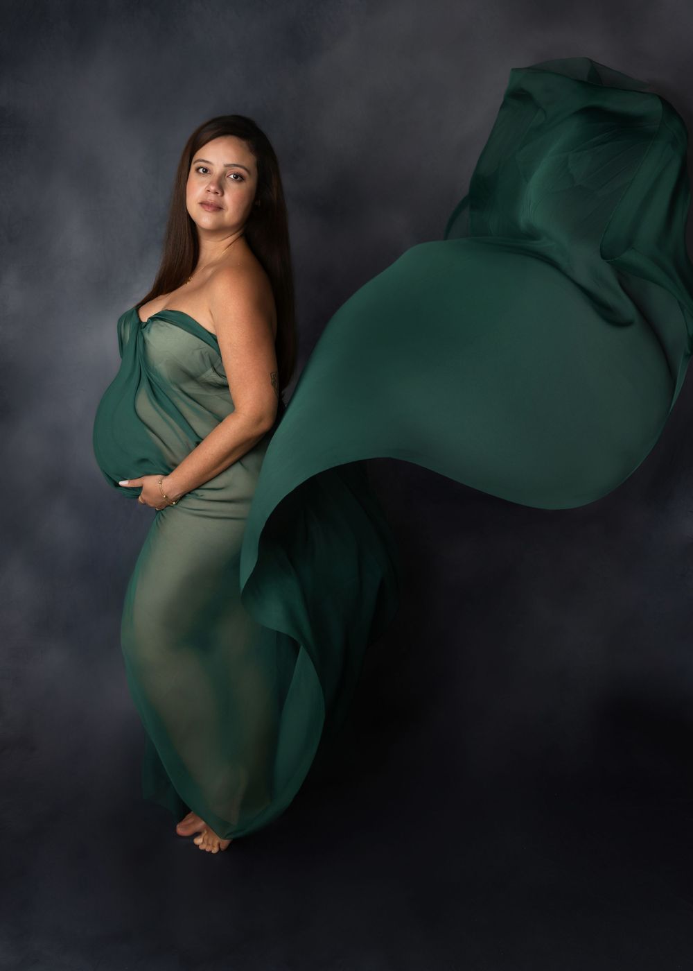 Pregnant woman in flowing emerald green gown with fabric billowing, posed elegantly against dark studio backdrop.