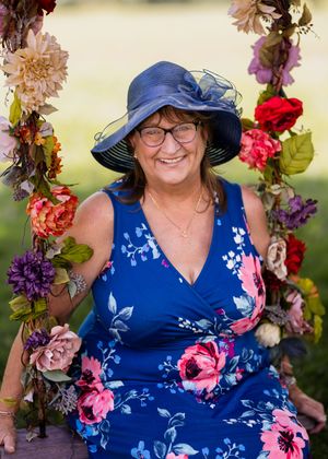Person in blue floral dress and navy hat poses among colorful flower arch.
