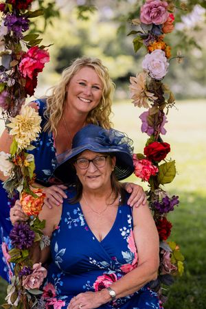 Two people pose together on a flower-adorned swing in matching blue floral dresses outdoors.