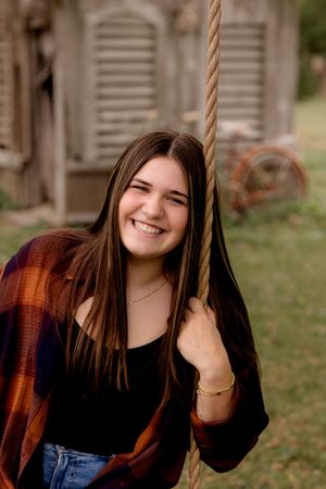 Portrait of someone smiling while holding a rope swing wearing plaid and denim against a rustic backdrop.