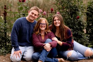Family of three sitting together outdoors with red roses in background during autumn photoshoot.