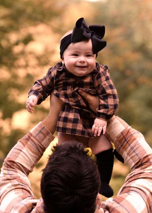 A baby wearing plaid and black bear ears is lifted playfully in the air against fall foliage.