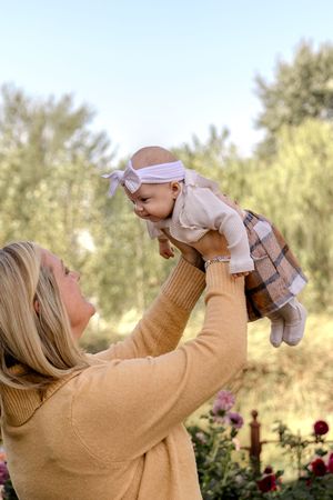 Person in beige sweater lifting baby against autumn outdoor backdrop.