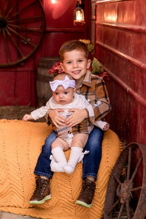 Baby sitting on orange crochet blanket in rustic red barn setting.