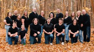 Large group wearing black clothes poses together in a long panoramic shot among fall trees.