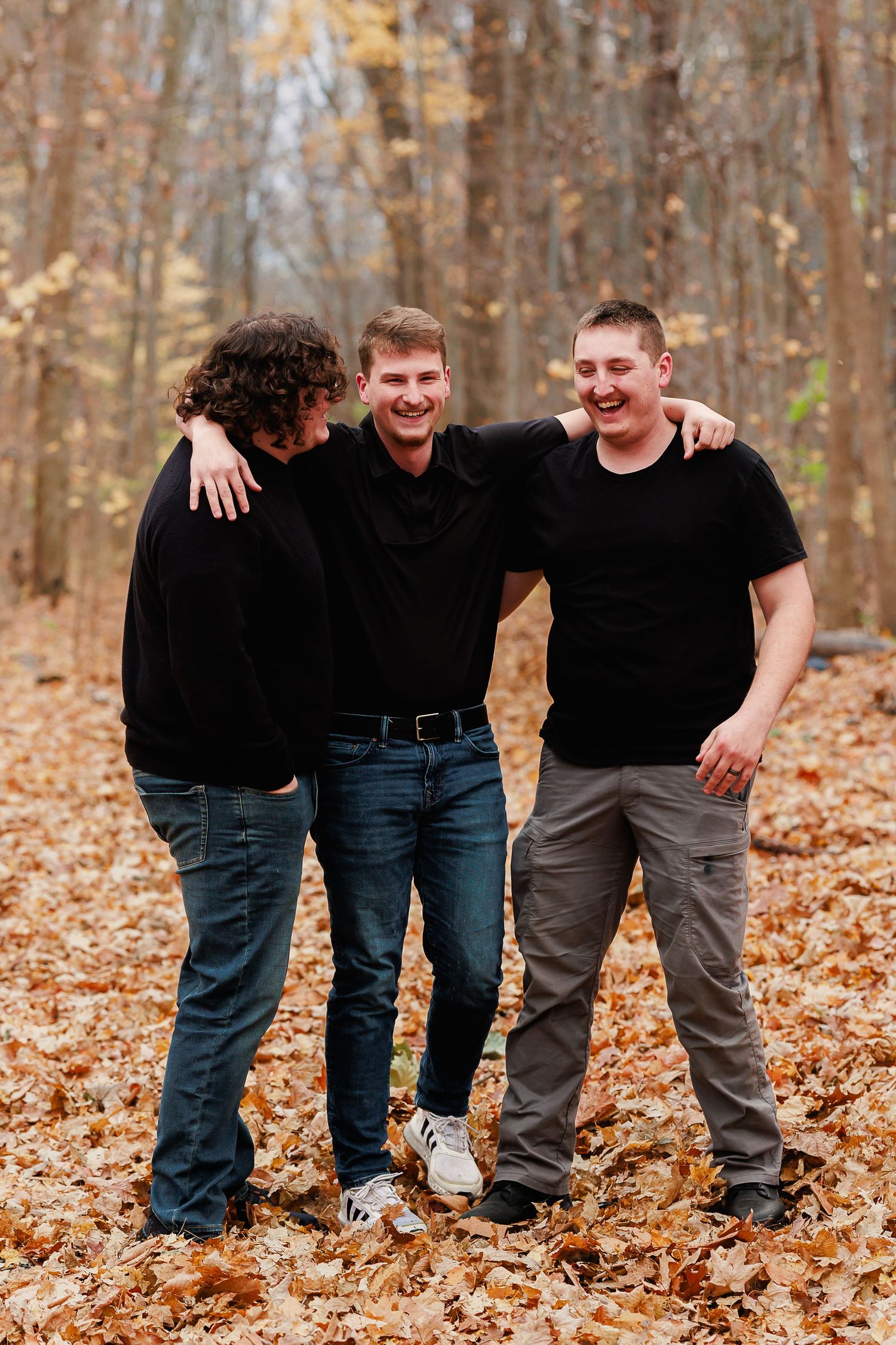Three friends in black shirts share laughs while standing in autumn woods.