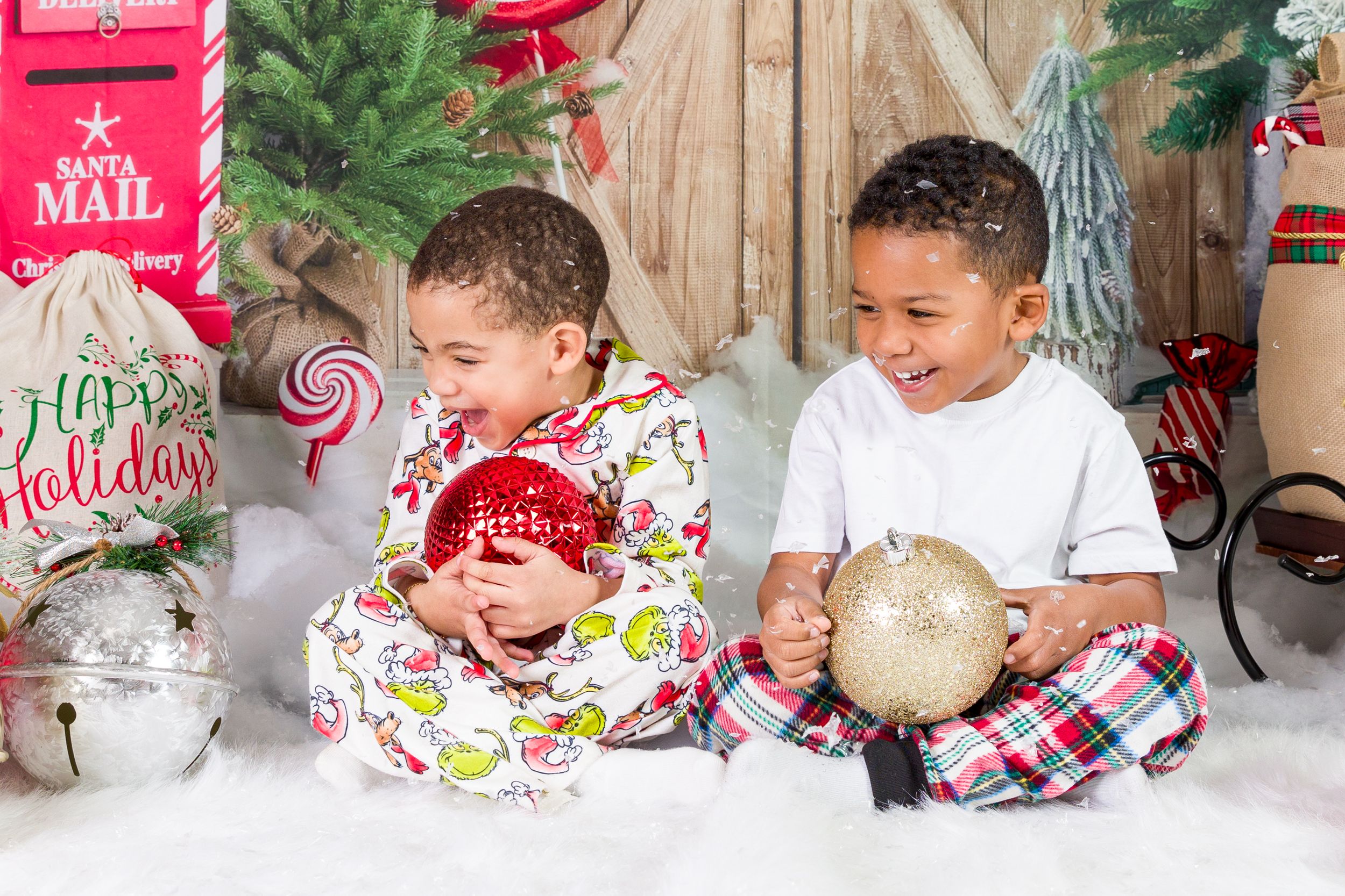 Two children in holiday attire sit on fluffy white carpet with Christmas decorations and a red mailbox in the background.