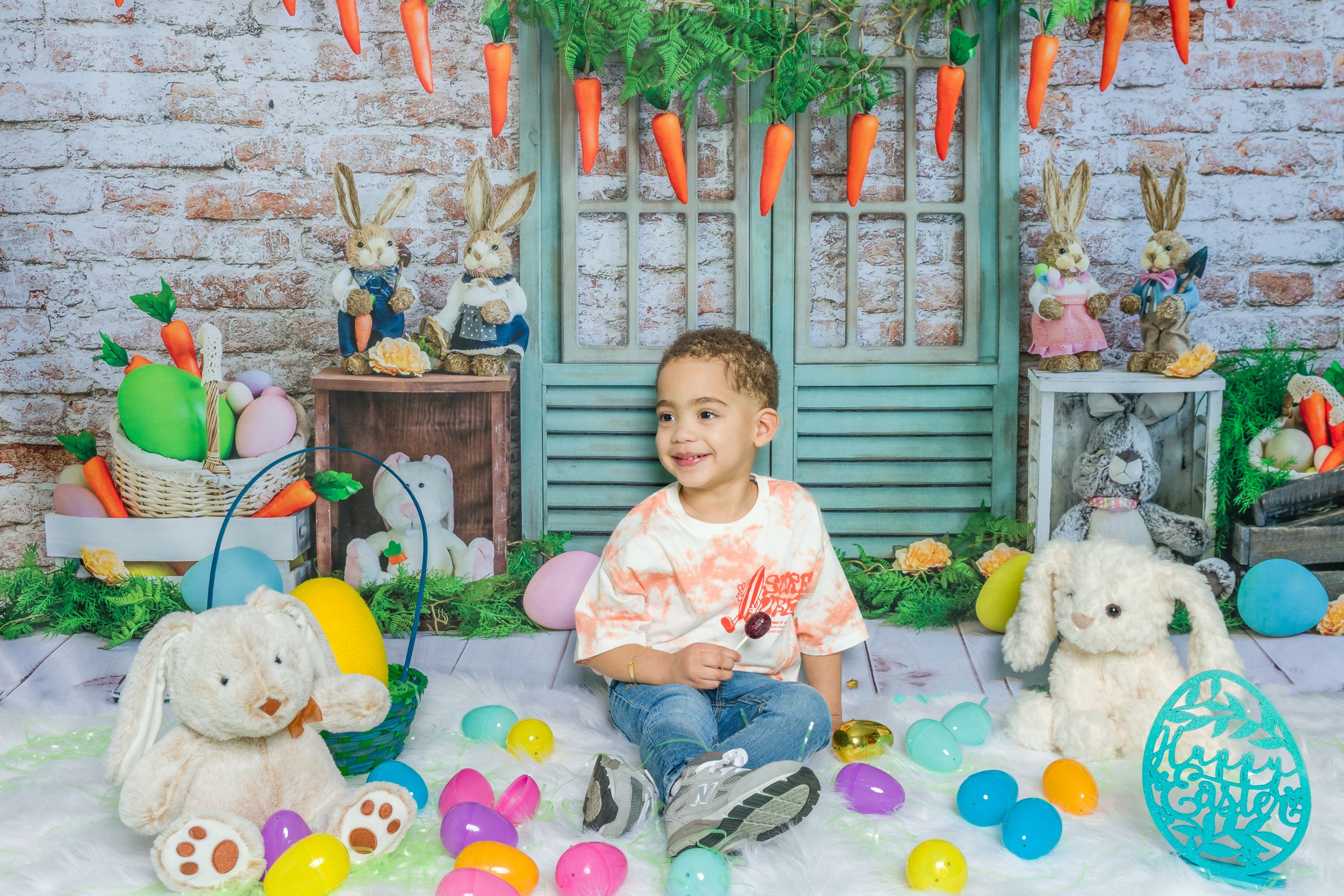 Easter-themed photo setup with colorful eggs, carrots, and stuffed bunnies against a rustic brick wall backdrop.