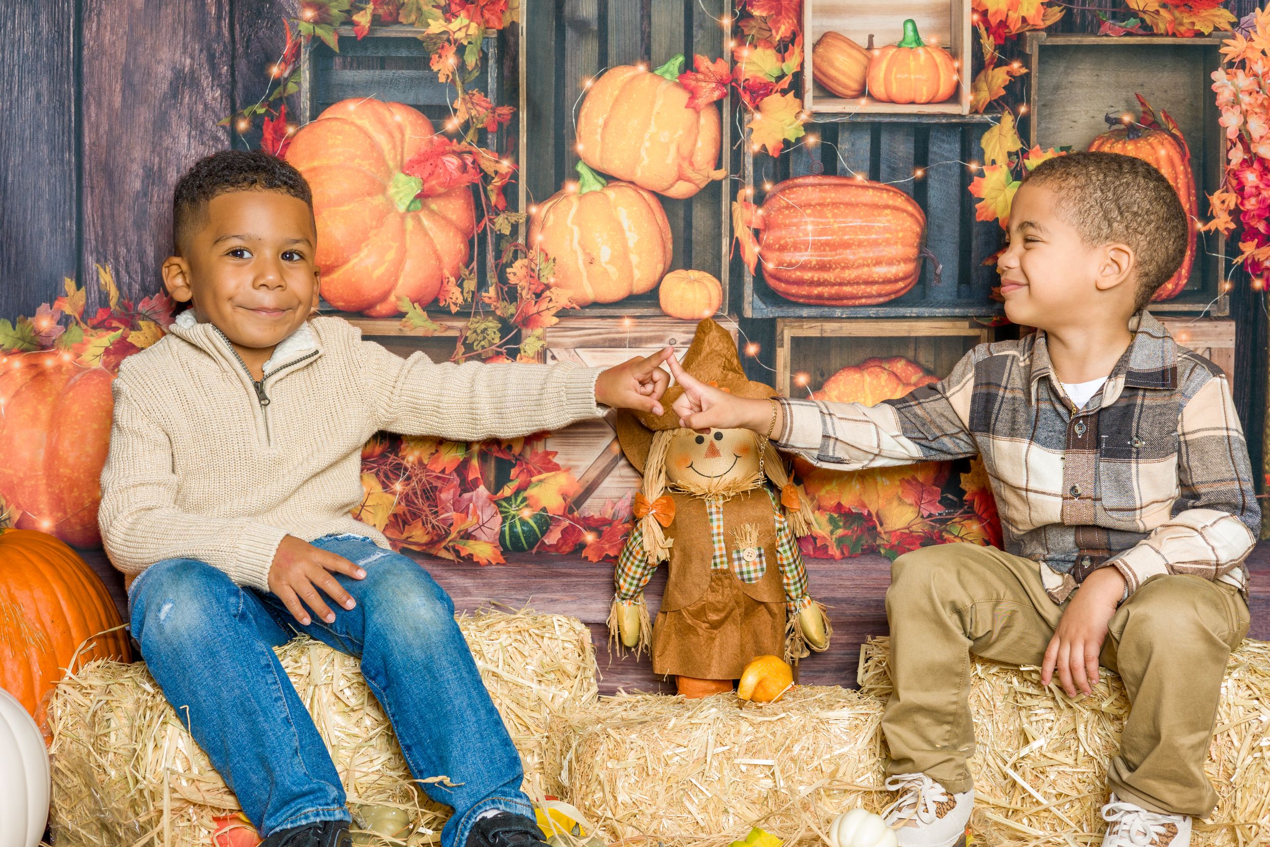 Two children sitting on hay bales with pumpkins and autumn decorations in rustic setting.
