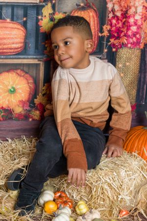 Child in striped sweater sitting on hay bale surrounded by autumn pumpkins and fall decor.