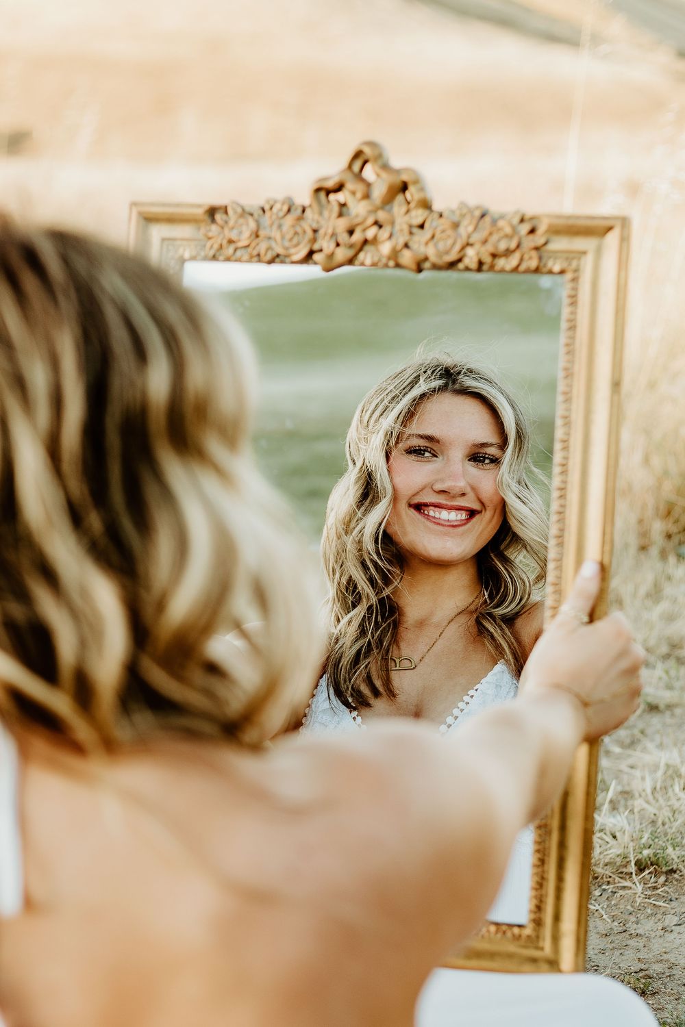 Young blonde woman smiling at her reflection in an ornate gold-framed mirror outdoors in a sunny field.