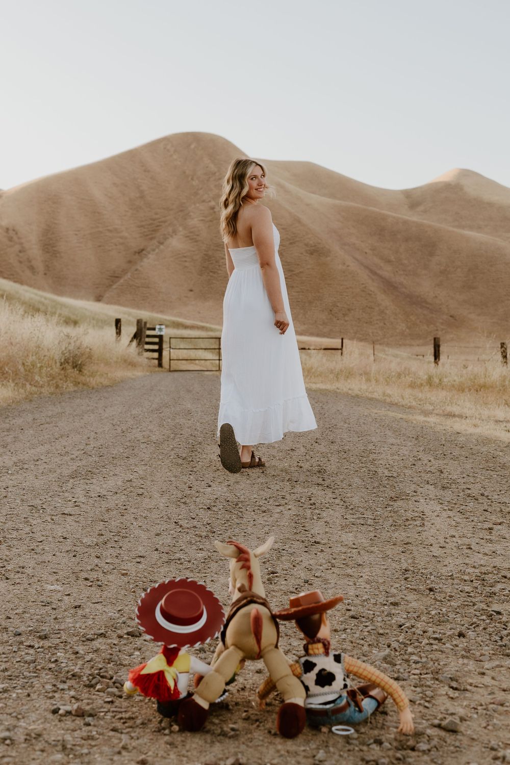 Woman in white dress on dirt road with Toy Story Woody and Jessie plush toys, golden hills in background.