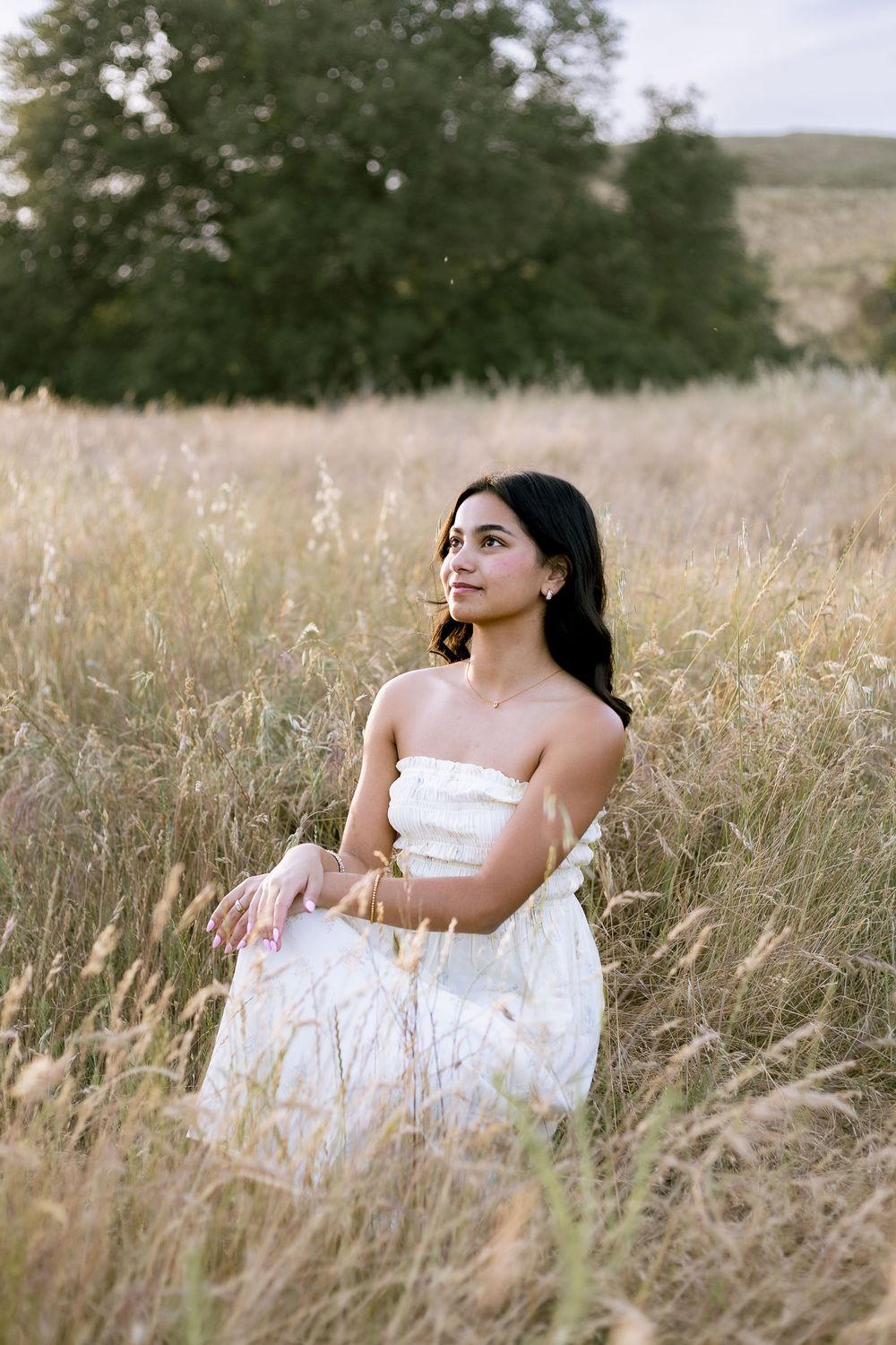 Young woman in white strapless dress sitting gracefully in golden field with trees in background.