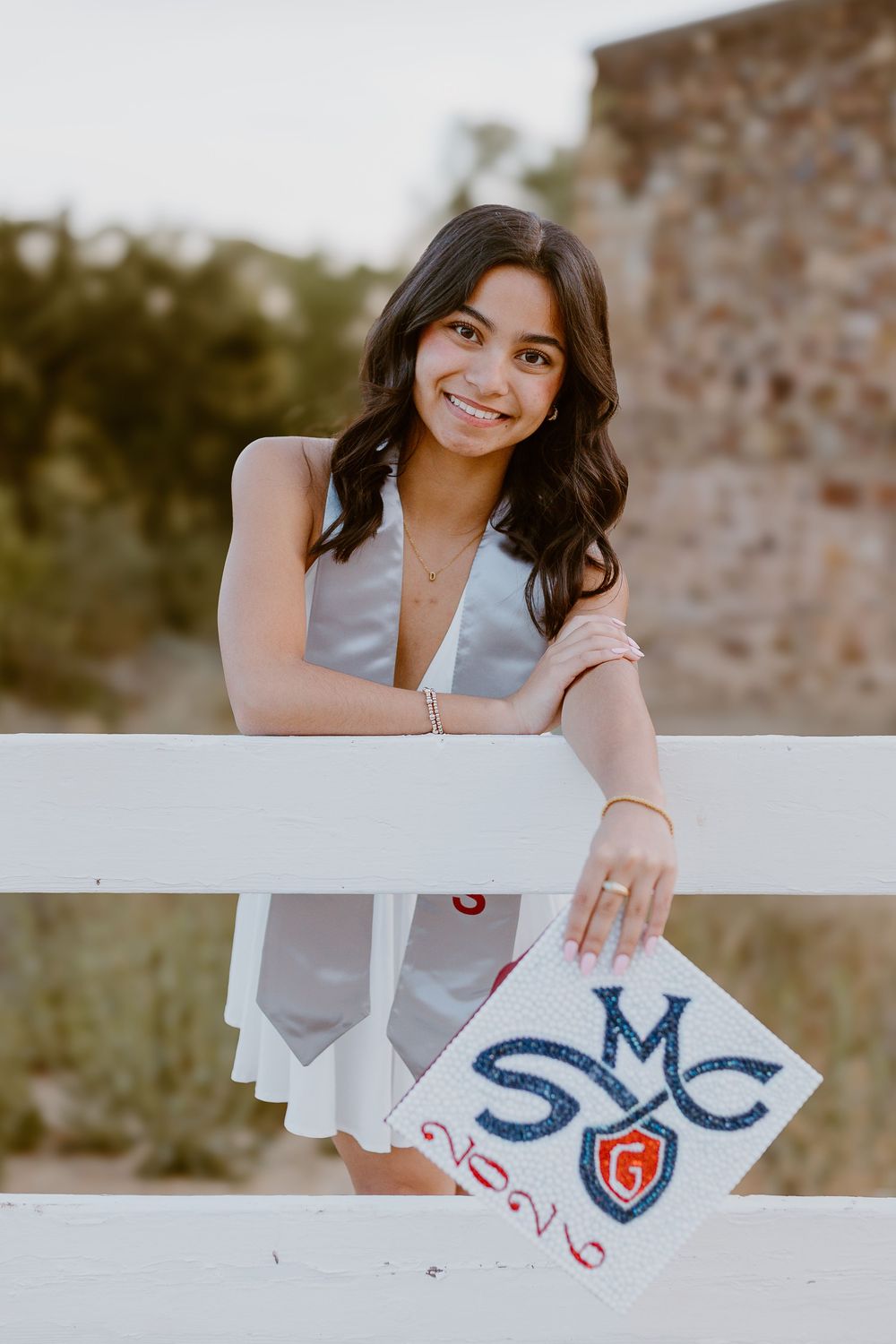 Smiling graduate in white dress holding decorated SMC graduation cap while leaning on white fence outdoors.
