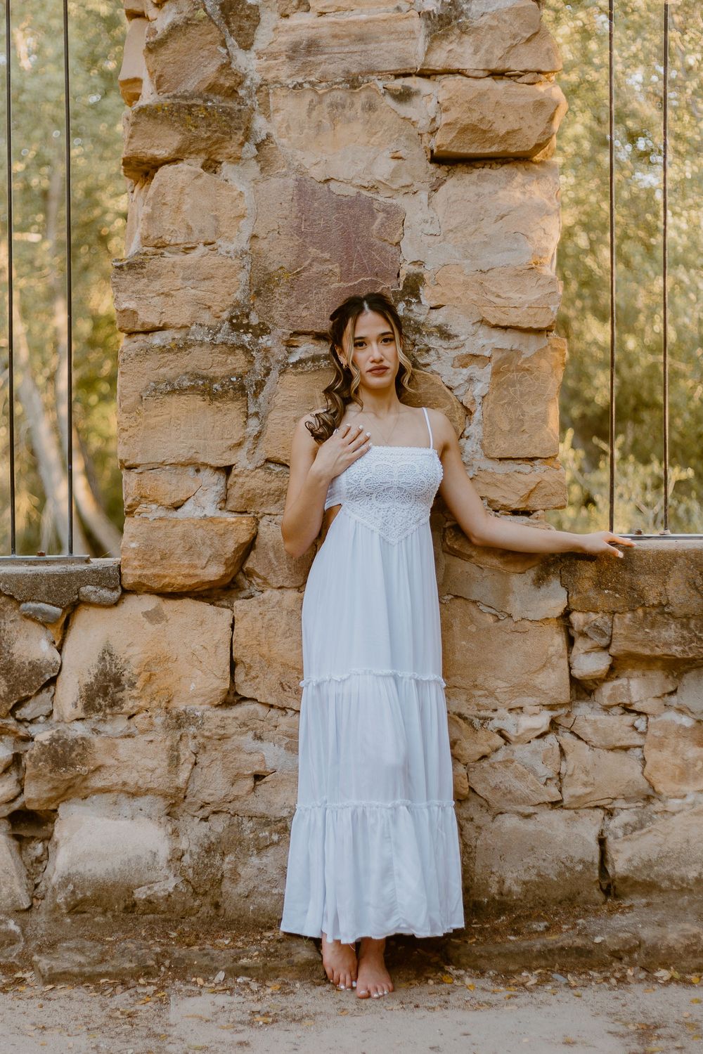 Young woman in white maxi dress posing barefoot against rustic stone wall outdoors.