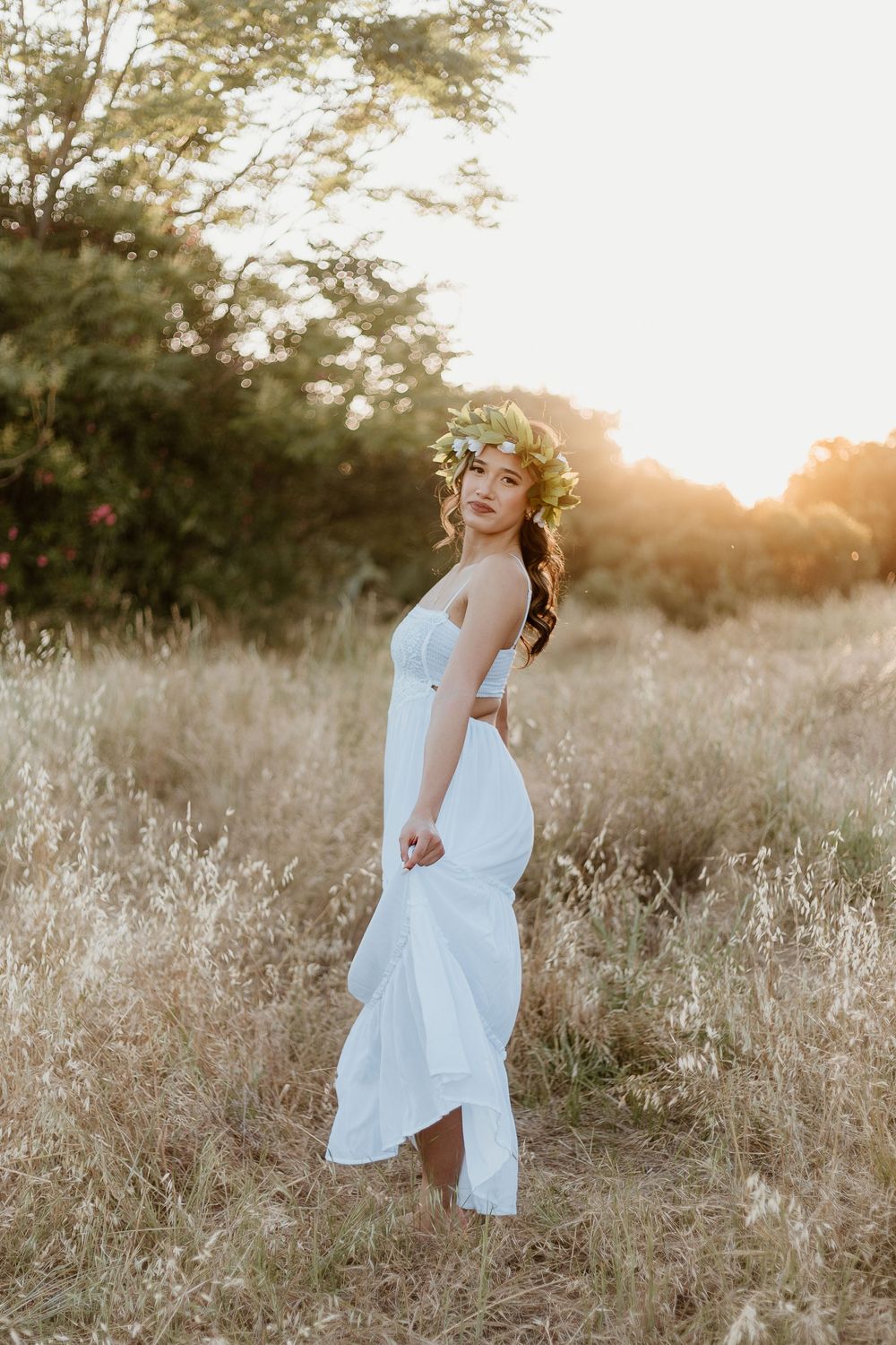 Young woman in white dress and floral crown stands in golden field at sunset.