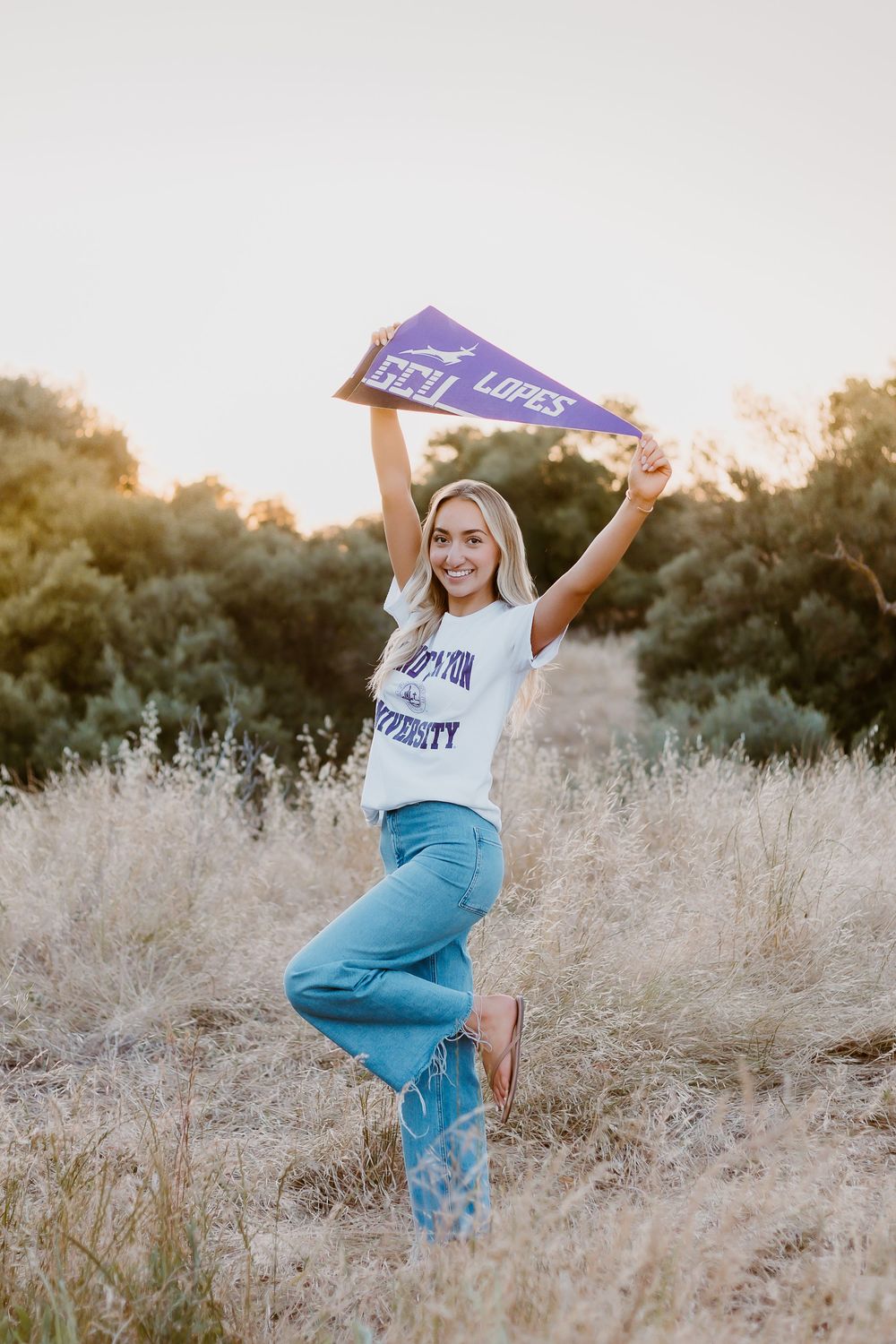 Young woman holding GCU Lopes pennant in a sunny field, wearing white tee and blue jeans.