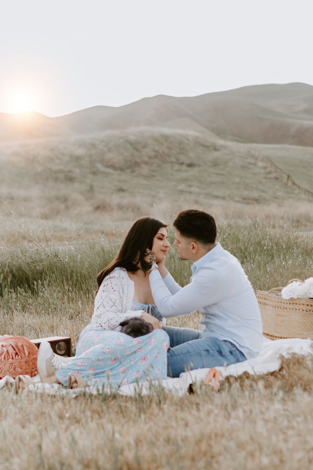 A couple shares an intimate moment during sunset on a picnic blanket in golden rolling hills.