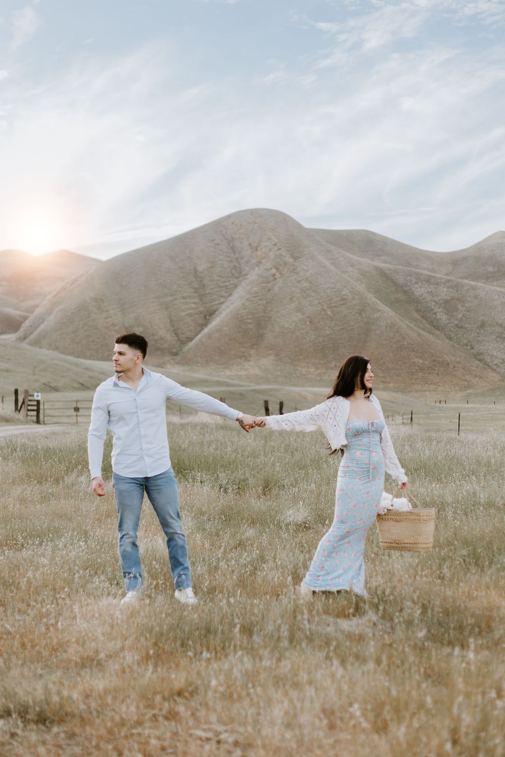 Couple in light colored clothing holding hands in a grassy field at sunset with rolling hills in the background.