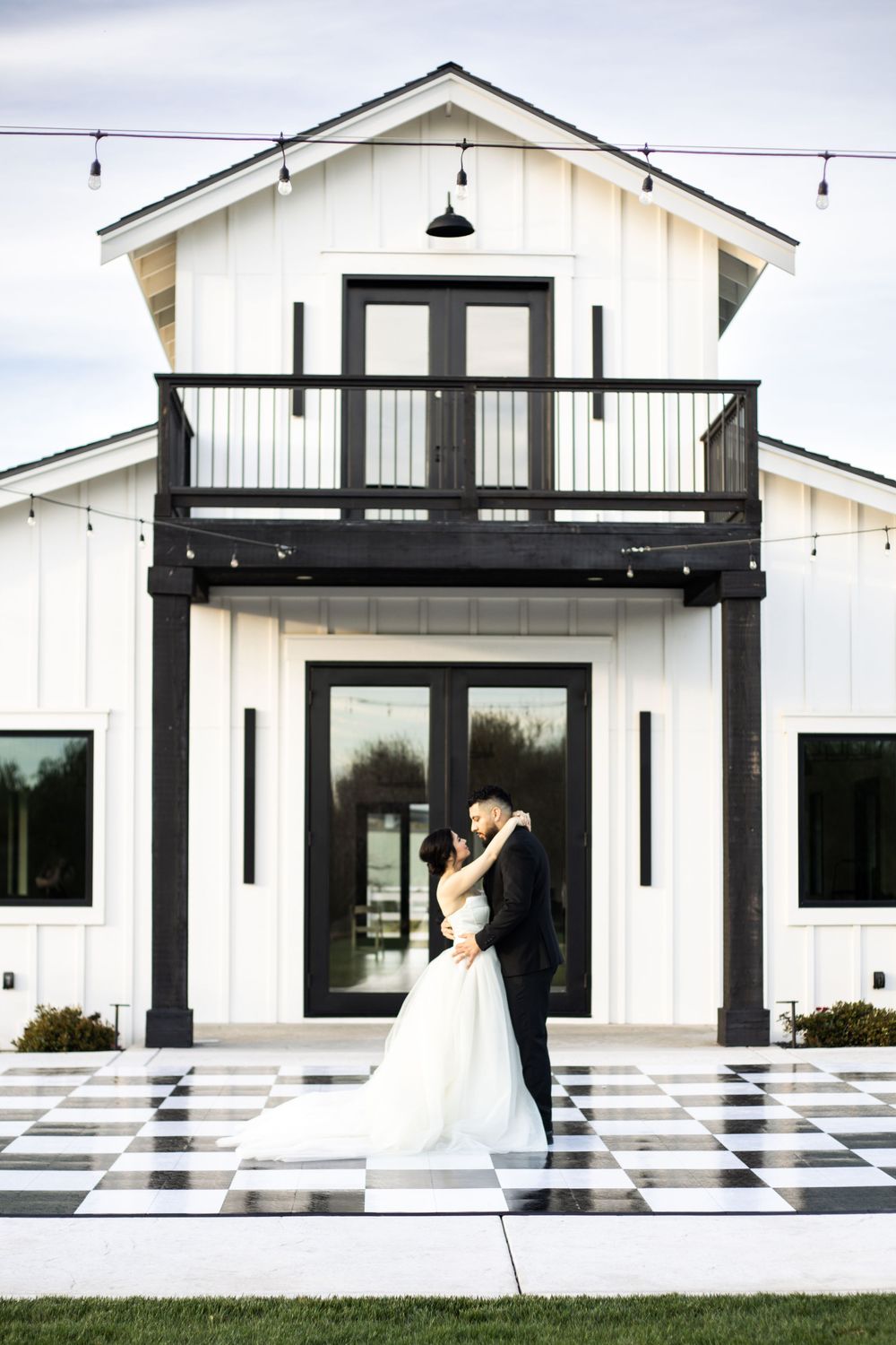 Bride and groom share a romantic kiss on a black and white checkered dance floor outside a modern white barn venue.
