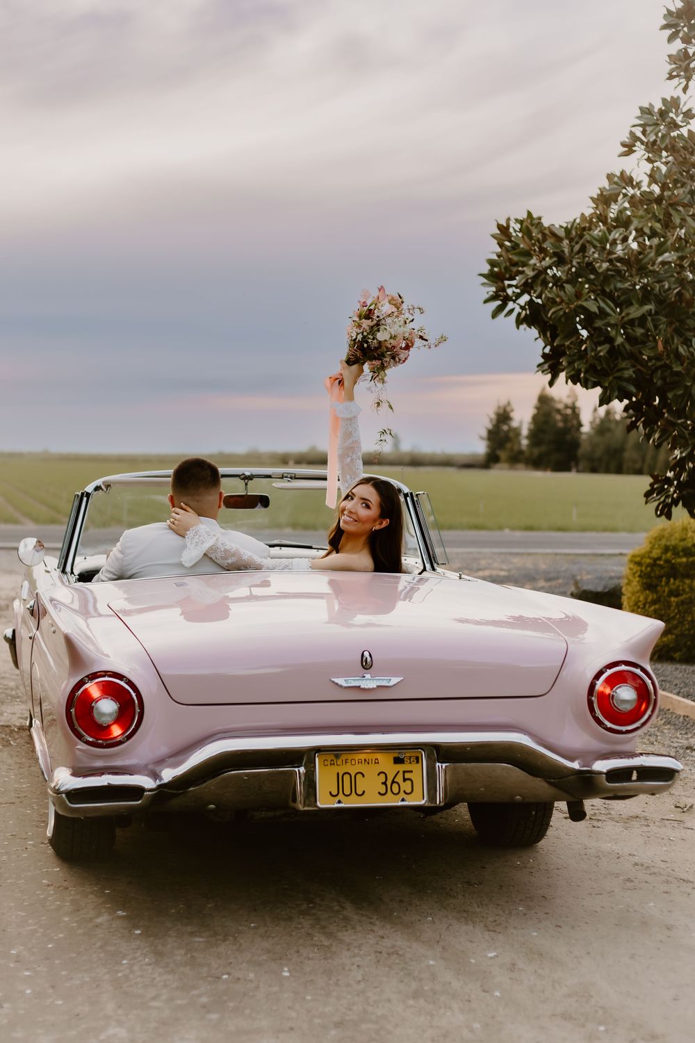 Bride raises bouquet in pink vintage Ford Thunderbird convertible with groom at sunset wedding celebration.