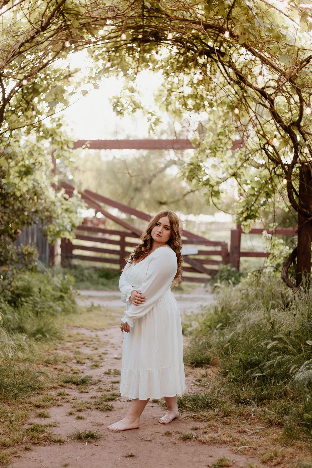 Pregnant woman in white dress standing under a floral garden arch on a dirt path surrounded by lush greenery.