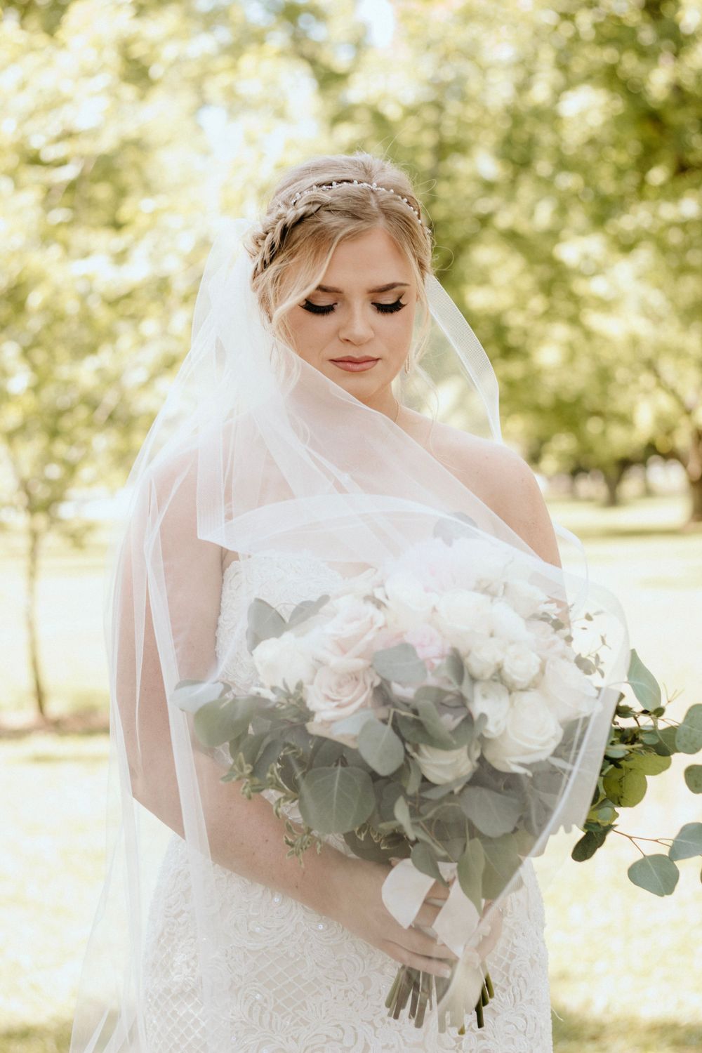 Elegant bride in white dress and veil holding lush bouquet with eucalyptus in sunlit garden.