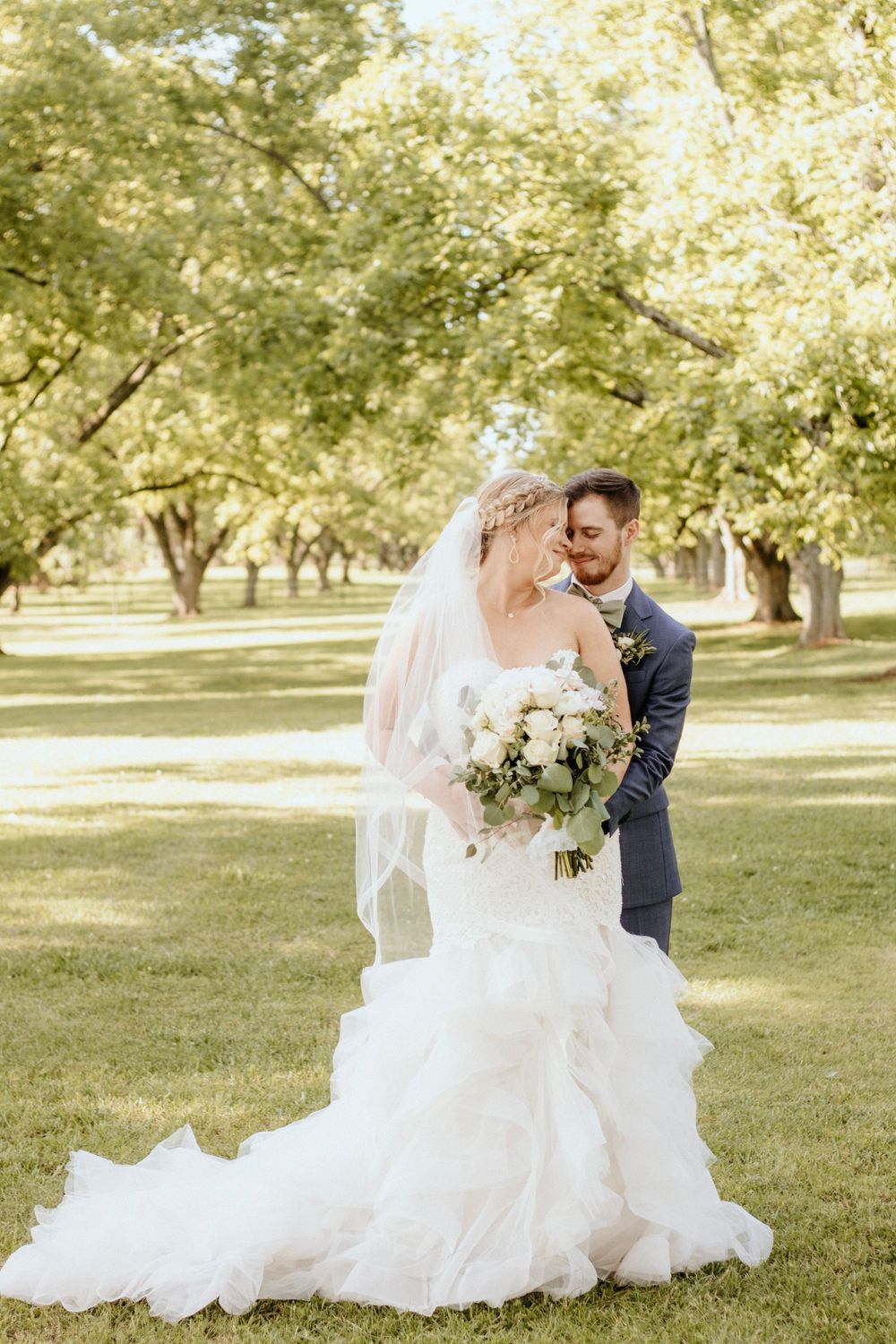 Romantic wedding portrait showcasing flowing white gown and classic navy suit in a sun-dappled garden.