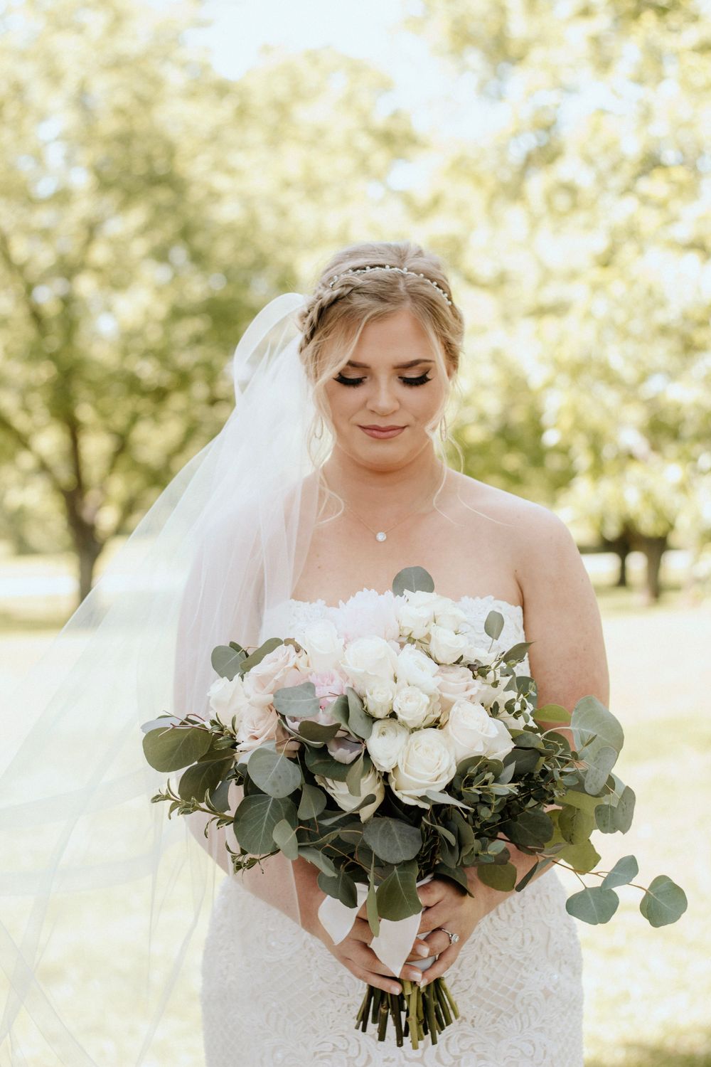 Bride holding romantic white and green bouquet in sunlit outdoor setting.