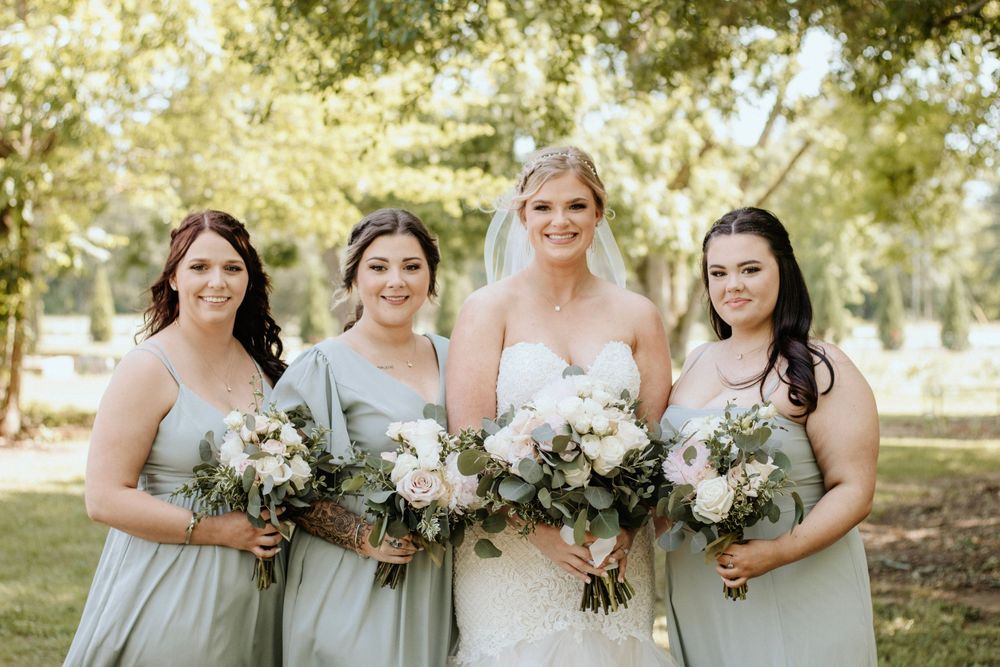 Wedding party in sage green dresses holding matching white floral bouquets outdoors.