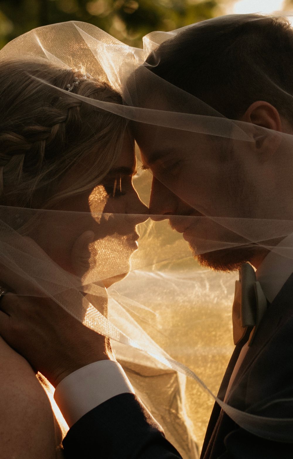 Artistic close-up of newlyweds kissing beneath flowing veil in golden sunlight.