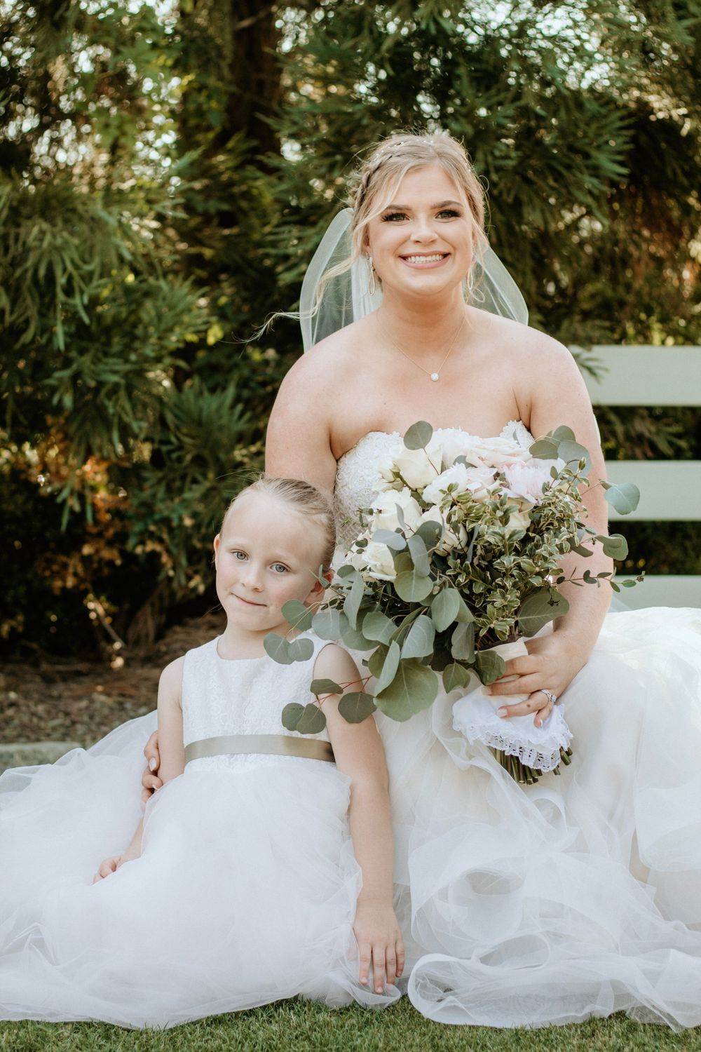 Bride in white wedding gown posing with young flower girl on garden lawn.