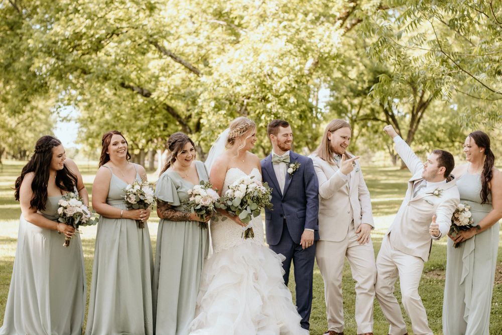 Wedding party in sage green and beige attire posing together in sunny garden setting.
