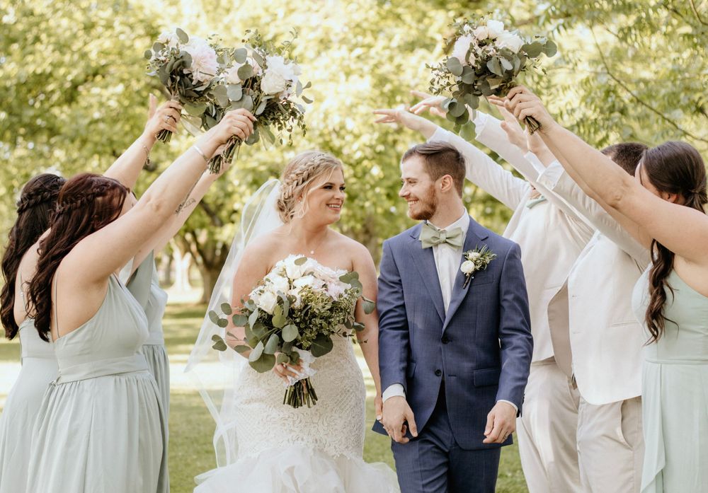 Wedding party celebrates with raised bouquets in an outdoor garden setting filled with natural sunlight.