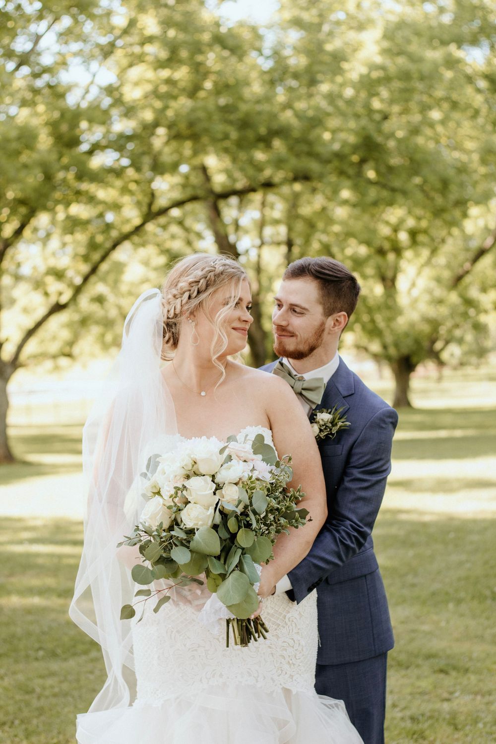 Elegant outdoor wedding photo featuring couple with white floral bouquet against sunlit trees.