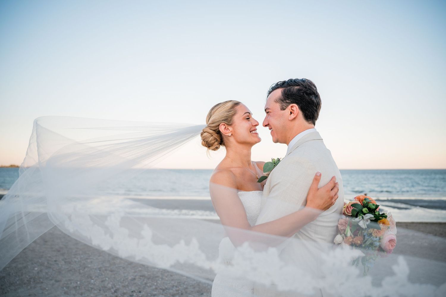 Bride and groom share romantic moment by the sea, veil flowing in the wind, holding a bouquet at sunset at Pavilion at Penfield Fairfield CT