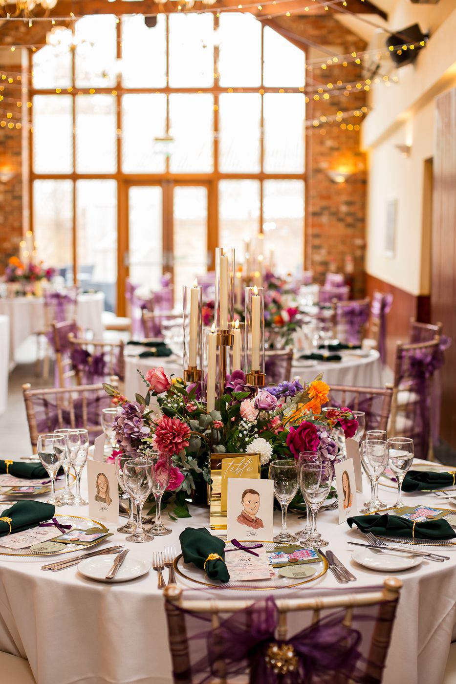 Elegant wedding reception table setting with colorful floral centerpiece and purple accents in rustic venue.