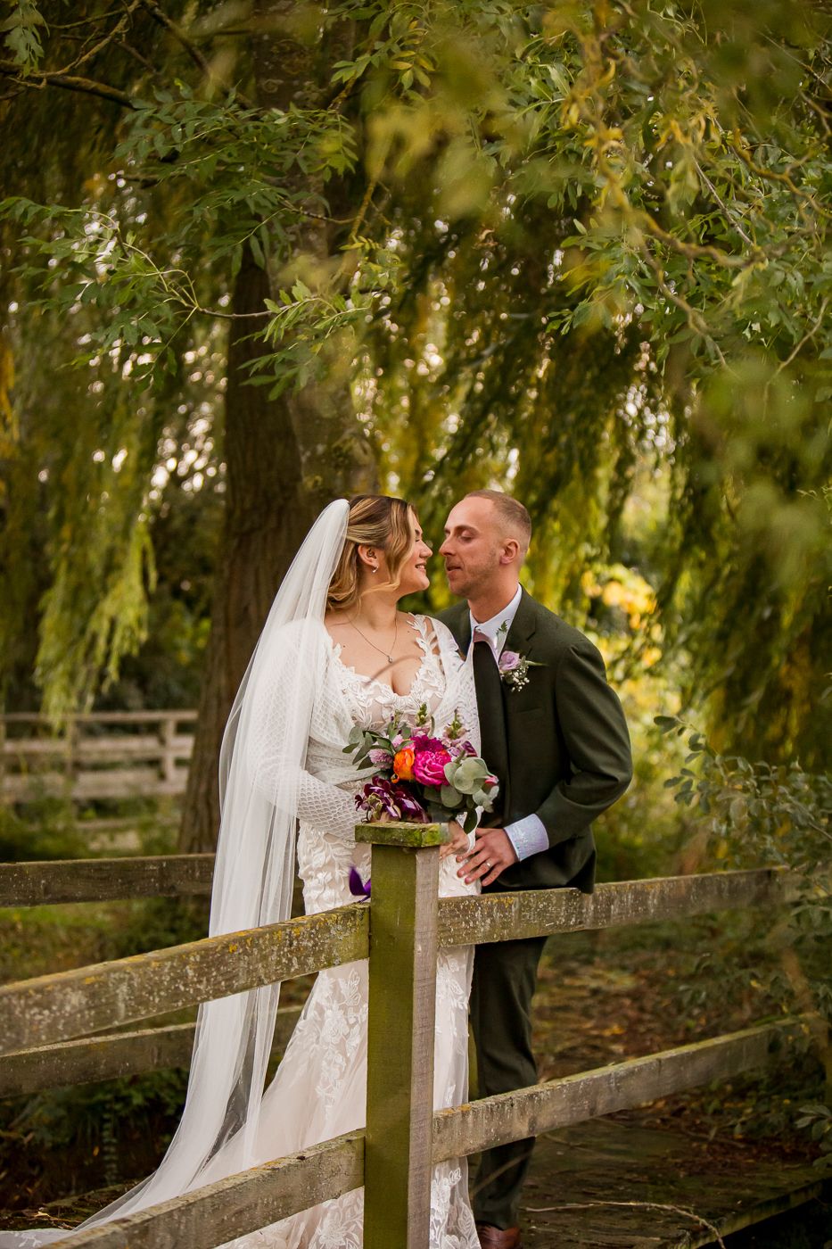 A romantic wedding moment captured on a rustic wooden bridge under weeping willow trees with a beautiful bouquet.