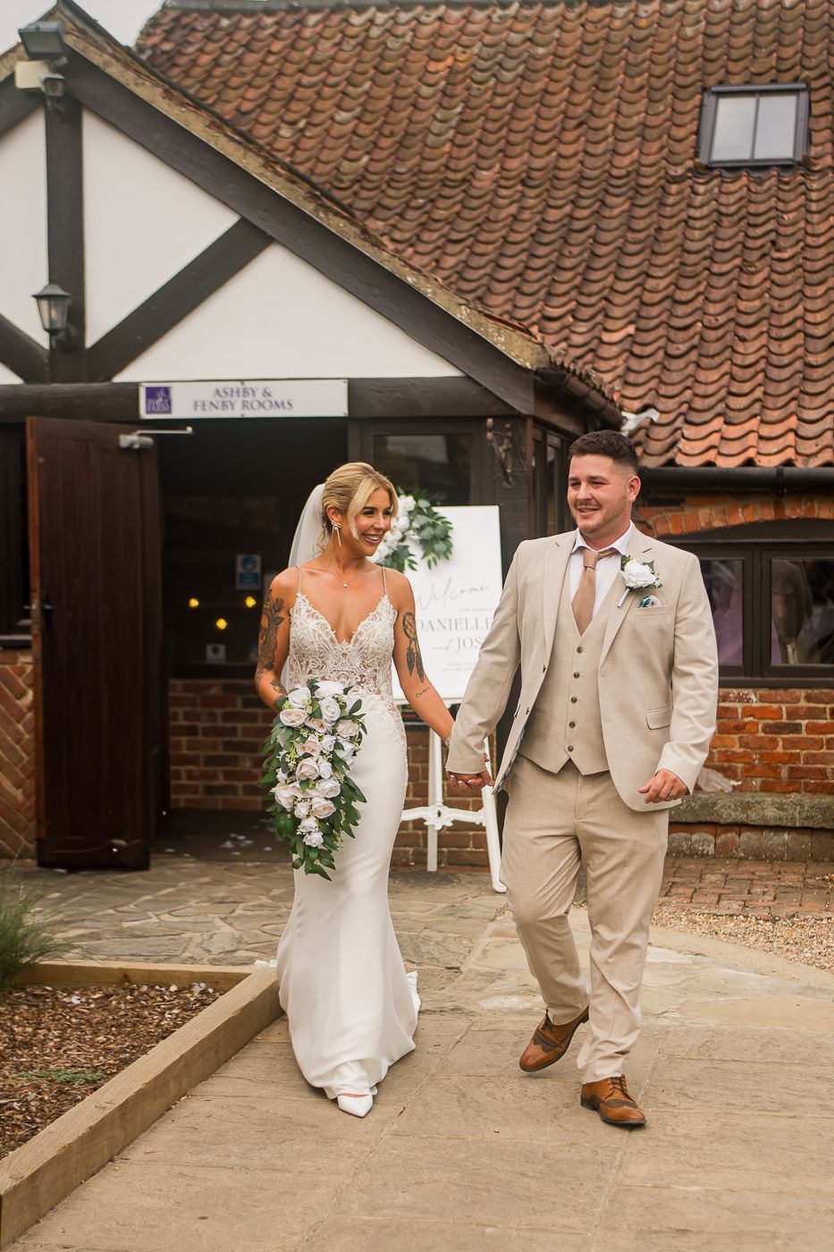 Couple walking together at rustic barn wedding venue with terracotta roof tiles and brick walls in background.