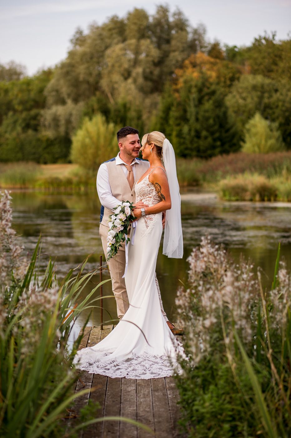 A romantic wedding couple stands on a wooden dock by a pond surrounded by tall grasses and autumn trees at sunset.